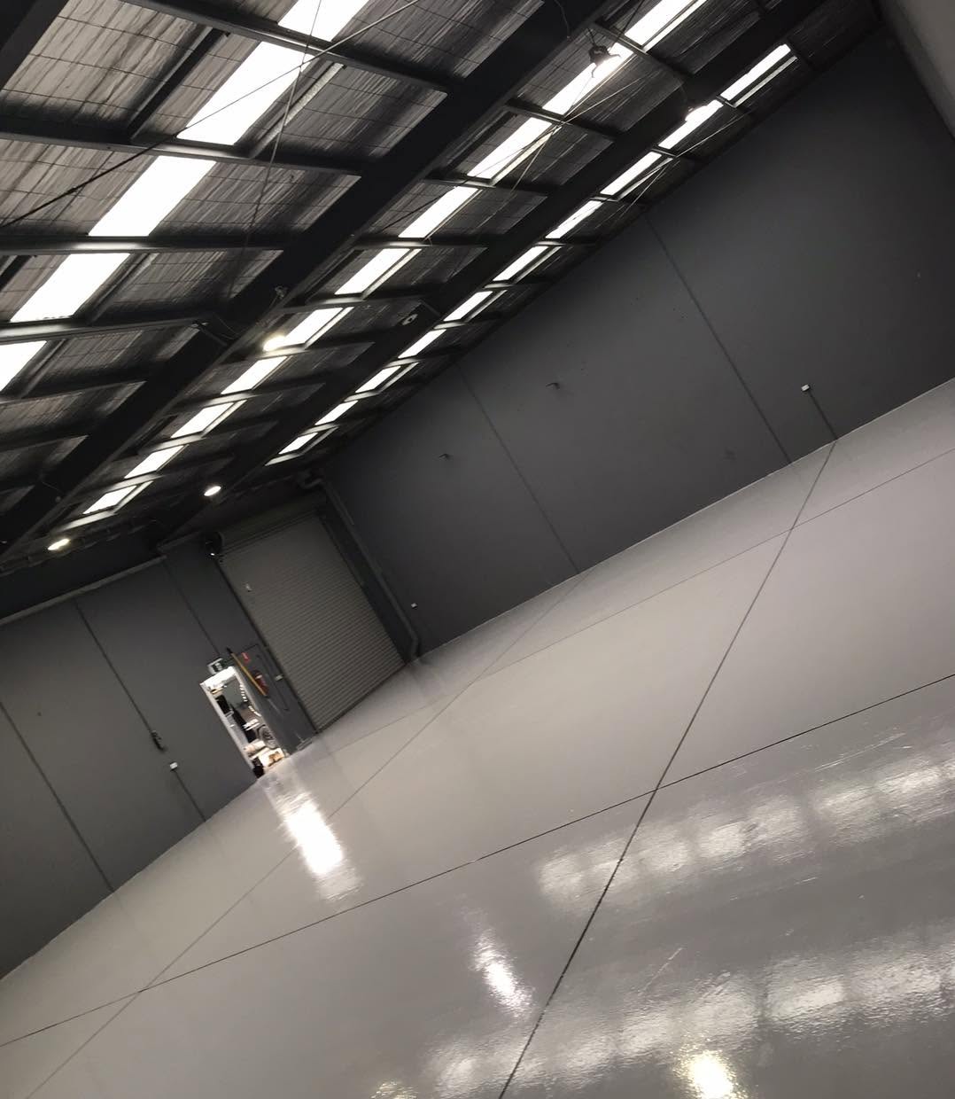 Long warehouse interior with a freshly sealed grey floor reflecting the roof skylights